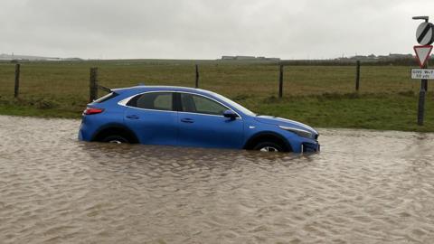 A blue car sitting in a big pool of water. Green fields in the background.