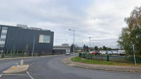 Google streetview image of a road, just coming off a junction, and curving round to the right. There is a car park on the right hand side and a large dark grey building on the left of the road. A brick building on the left is the police station.