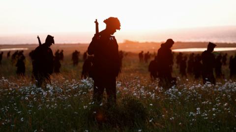 silhouettes representing British soldiers who died during World War II standing in a field of daisies with their heads bowed and dimming sunlight in background 
