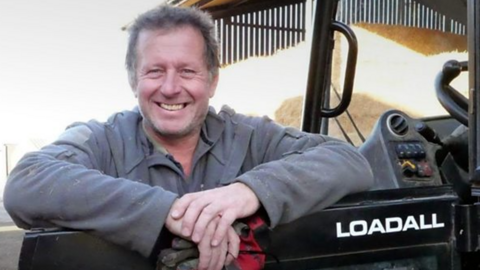 A man with short brown hair and a grey jacket, leaning one arm on the door of a black tractor cab with the word Loadall written in white writing on it