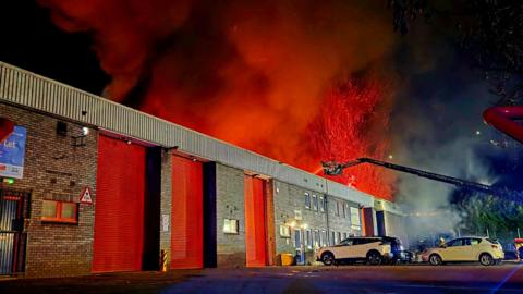 A large warehouse building on fire with three cars parked outside. There are huge red flames coming from the top of the building while firefighters on a cherry picker shoot water down