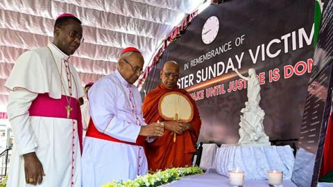 The head of Sri Lanka's Roman Catholic church Cardinal Malcolm Ranjith (C) places a candle in front of a plaque as he pays respect to the victims of the 2019 Easter Sunday bombings on the 6th anniversary of the suicide attacks, as Vatican ambassador to Colombo Archbishop Brian Udaigwe (L) and Buddhist monk Omalpe Sobitha watch outside the St. Anthony's church in Colombo on April 21, 2025