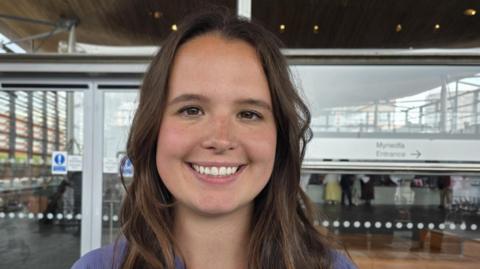 Zaynub Akbar smiling outside the Senedd in Cardiff  Bay with glass entrance door and windows behind her. She has long brown hair and smiles and wears a mauve shirt