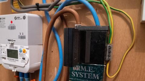 A black box attached to a chipboard wall, with electrical wires at either side and a electricity meter to the left