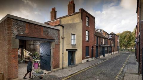The red brick Threadgolds building alongside a narrow paved street in Portsmouth. The nearest section has a flat roof and open doors with someone walking out pushing a wheelbarrow
