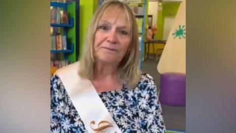 The image shows a person sitting in a colourful children’s library area, wearing a patterned top and a sash draped across the shoulder. Behind the person are bright green walls, blue bookshelves filled with children’s books, and soft play-style seating in purple and light colours, indicating a space designed for young readers.