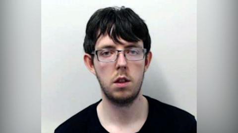 Police mugshot of a pale man with black messy hair pictured against a white background. He is staring directly at the camera wearing a black t-shirt. He has heavy dark stubble and is wearing glasses.