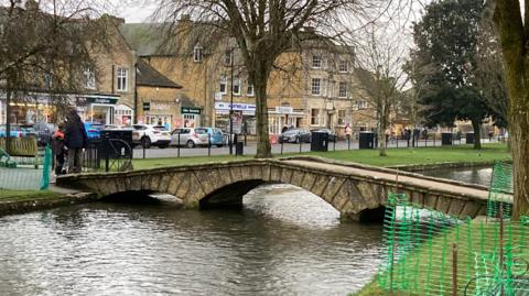 A small stone footbridge with three arches over the River Windrush. The water comes right up to the banks. Behind the bridge is grass, with a few trees and behind that is a row of shops, with cars parked in front.  