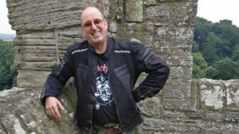 A bald man is leaning on an old wall and smiling at the camera. He is wearing sunglasses, a black motorbike jacket and a Motorhead T-shirt.