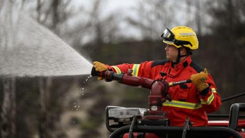 A Surrey firefighter in red and yellow firefighting kit manning a water hose