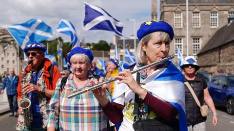 A woman draped in a saltire plays a flute at the front of a crowd of pro-independence marchers