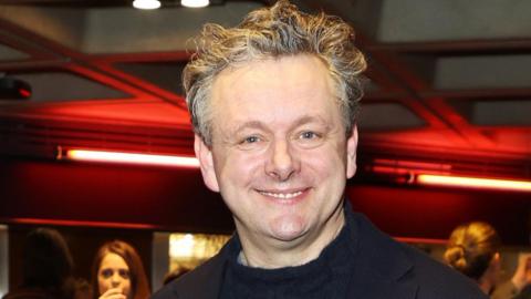 Picture of Michael Sheen at a Nye musical launch event. He has silver wavy hair and smiles widely at the camera. He wears a black funnel neck jumper and a black collar can be seen. 