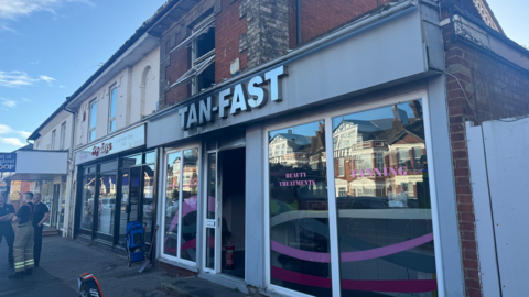 A photo from the pavement of Tan-Fast tanning salon. Firefighters are standing outside of the building to the left of the image.