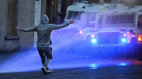 Rioters are targeted by a PSNI water canon during a third night of civil unrest in Ballymena. A rioter can be seen in a grey hooded top, grey trousers and grey trainers - standing in front of PSNI vans in the street with both his arms raised at them. Water from the cannons can be seen splashing at him. The water is partly illuminated by the blue lights from the police van.