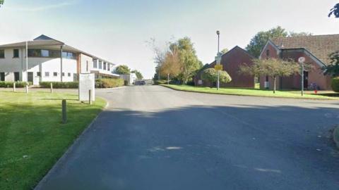 The entrance to the Newton Rigg College campus, with several buildings on either side of the road, surrounded by patches of grass and trees. The building on the left of the central road is mainly white and two storeys high. On the other side are red brick buildings that resemble houses, with roofs which slope down further on one side than the other. The area is empty of people and vehicles.