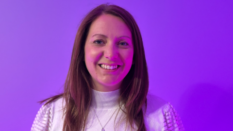 A woman with long brown hair and a white top in front of a violet background, lit with a violet light