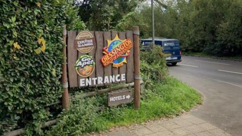 Chessington World of Adventures car park entrance with brown sign showing Chessington, Go Ape and Explorer Camping logos on them and a smaller arrow sign for the hotels.