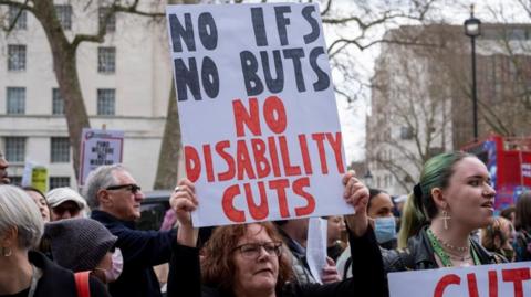 A female protestor in a crowd of people holds up a sign which reads "No disability cuts".