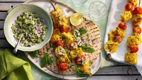 An oval platter topped with flatbreads and skewers of marinated cooked chicken with a bowl of green salad on a wooden outdoor table