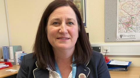 A woman with dark hair wearing a navy blazer sits in an office room, behind her on the wall is a map of Shropshire