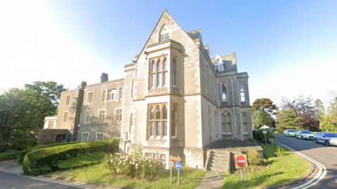 A Grade II* listed Victorian gothic mansion stands in gardens. The building features ornate arched windows.