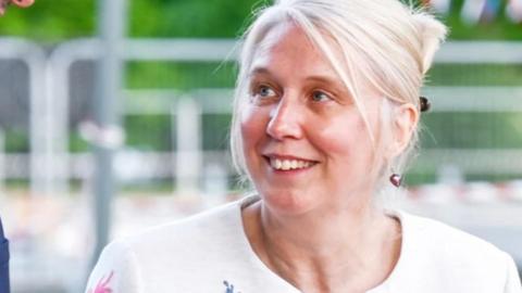 A woman with blonde hair tied up behind her head and a white top, smiling and looking away to her right