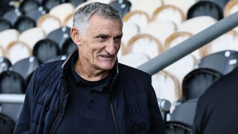Tony Mowbray is wearing a dark shirt and jacket, and there are empty stadium seats in the background.