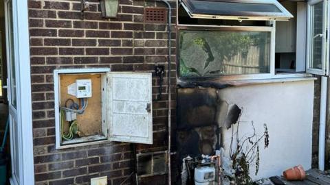 A burnt section of a wall beneath a window. There is a gas meter nearby and another meter on the ground beneath the burnt area