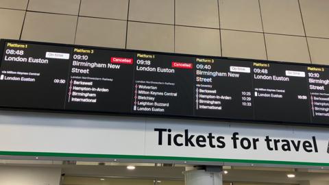 A train station with an information board with train times on it, showing cancellations and information. The screen is black and white.