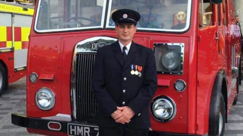 Man in smart black suit with a hat and several badges on his lapel. An old red fire truck is behind him and he stands with his arms clasped.