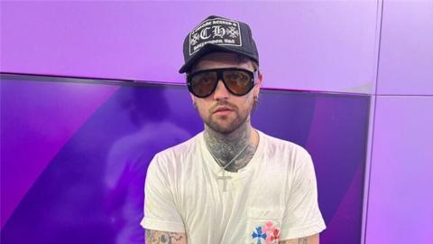 Mason Newman is stood in front of a purple background wearing shades,a cap, white T-shirt and silver chain.