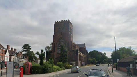 A large brown brick abbey with a road on the right hand side, and terraced buildings on the left. The sky is grey and cloudy
