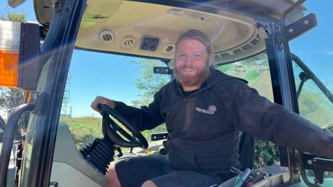 A picture of James sat inside a vehicle. He has a ginger beard and is wearing a black jumper.
