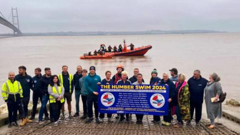 Twenty men and women stood in front of the River Humber on pavement holding a blue and yellow sign reading 'The Humber Swim 2024'. Most of them are dressed in blue uniform and some of them have hi-vis vests on. In the background are two Humber Rescue orange lifeboats with volunteers on board. 