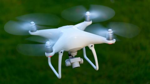 A white drone with four propellors and a camera looking down underneath it