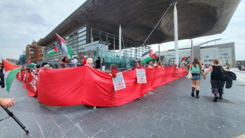 Protesters hold long red cloth wrap around the Senedd in Cardiff Bay. A couple turned away from the camera are holding hands at the right of the shot. The red banner can be seen going off to the left hand side.