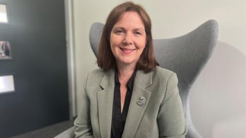 Woman with short brown hair in green linen jacket with black blouse and silver broach sits in grey armchair 