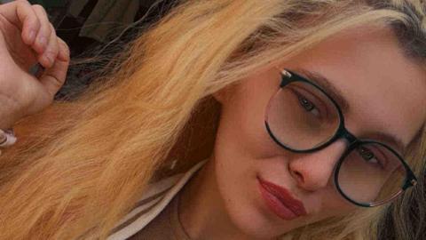 A young woman with glasses has long, wavy hair dyed blonde. One hand is raised near her head, and the background shows a dimly lit room.