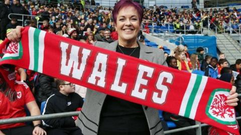 Dawn Bowden, who has short purple hair and is wearing a black and white jacket and black top, holding up a red Wales football scarf and standing in the stands of a sports stadium, with swarms of other spectators in the seats behind her