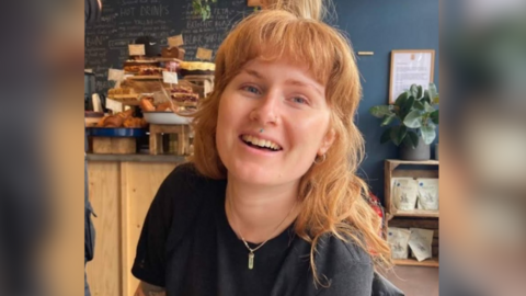Ruby Love, wearing a black t shirt and necklace, smiling at the camera. She also has a septum piercing and wears her long strawberry blonde hair down with a fringe. Behind her are what looks like cake and displays that show a cafe. 