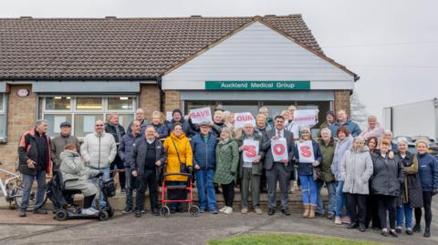 About 30 people are standing outside St Helens surgery owned by the Auckland Medical Group. Some of them are holding pieces of paper which say Save our Surgery, another three are holding the letters SOS up. 