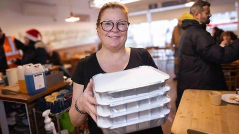 Kath Coogan wearing black glasses and a black t shirt holding four trays of takeaway food, smiling at the camera. She is stood behind the counter of a cafe, with people scattered in the background. 