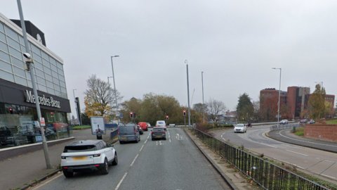 Traffic drives towards the city ring road from the Penn Road. It passes a Mercedes Benz car garage on the left with traffic lights at the island.A railing separates the traffic coming in the opposoite direction.