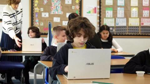 School children sitting at desks in a classroom setting with laptops.