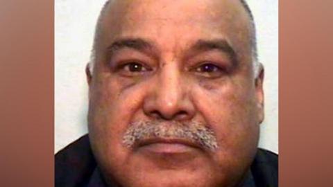 A mugshot of an Asian man in his 60s, with a grey moustache and bald head. He is looking into the camera.
