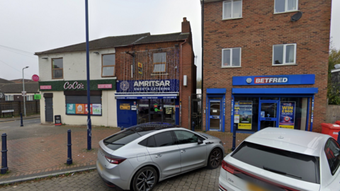 The shop is pictured in between two others and has blue sign outside with decorative lights cascading from near the roof down to above the front entrance 
