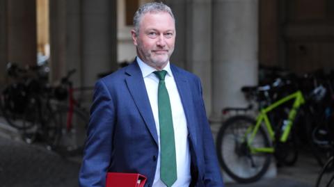 Steve Reed looks at the camera as wearing a navy suit with white shirt and green tie. He is carrying a red folder. He is walking through a passageway with bikes parked in the background.