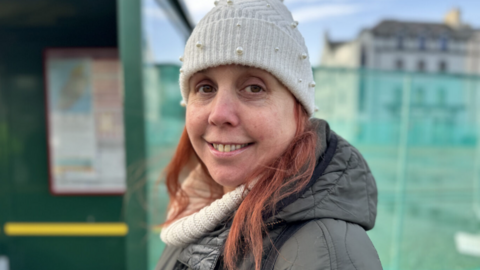 Kim Hidson is smiling wearing a white woolly hat with her red hair at the sides. She is wearing a woolly jumper and waterproof coat and is standing in front of a green bus shelter.