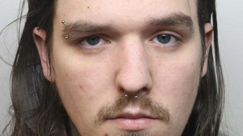 A police mugshot shows a man with long brown hair, a moustache and a goatee style beard, along with piercings in his nose and his right eyebrow.