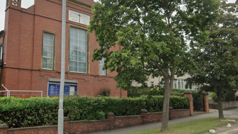 A red brick building with a blue sign at the front with the name of the church. A tree and hedgerow can be seen around the church.